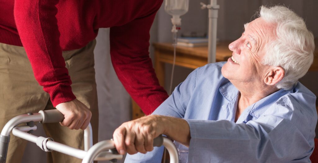 Physiotherapist assisting older man who is sitting down with his walker to stand up.