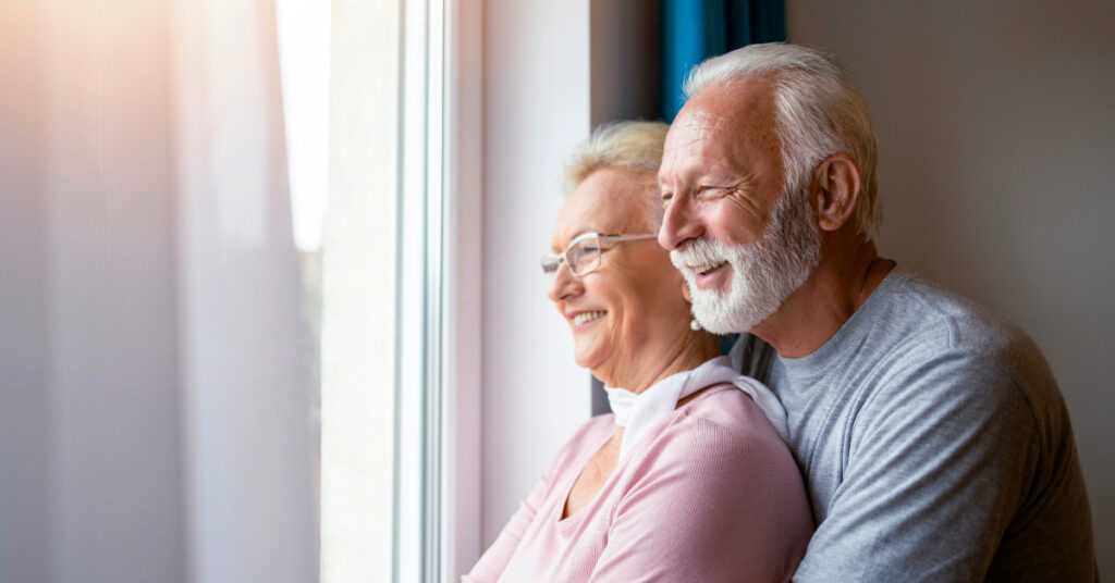 Healthy older couple in loving embrace, looking out the window.